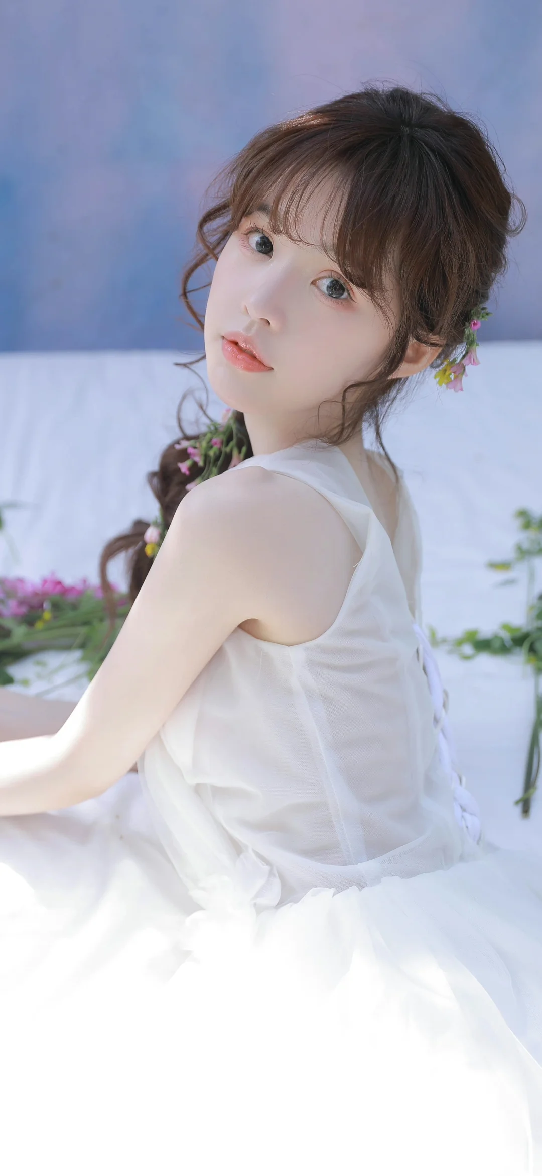 一名身穿白色无袖连衣裙的女性回头望向镜头，背景为浅蓝白色布景和少量花卉。