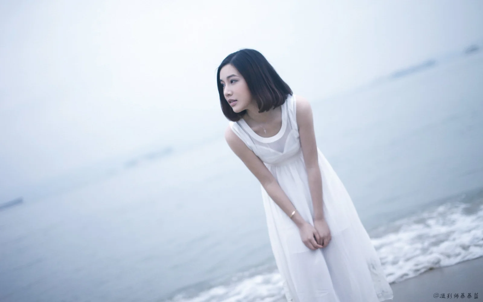 一名身穿白色連衣裙的女子站在海邊，身後是柔和的海水與浪花。