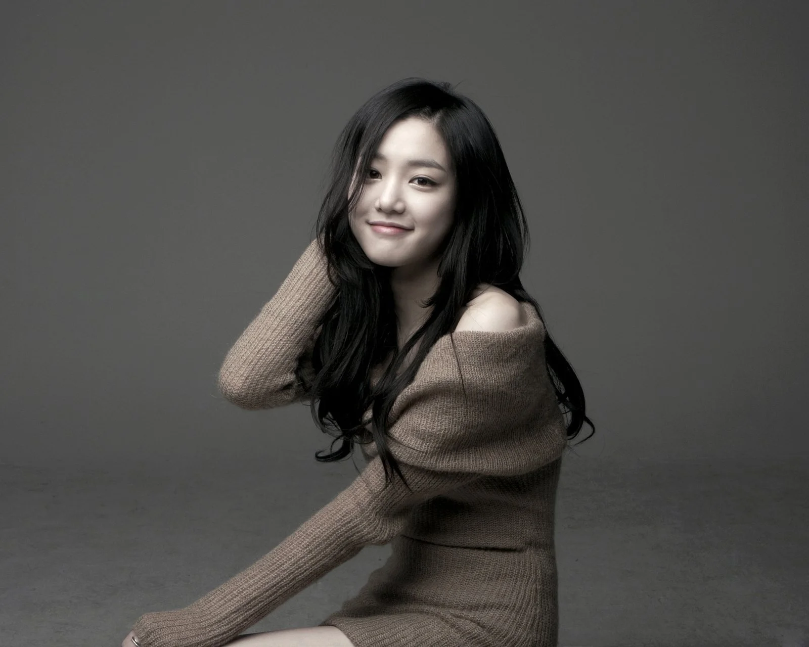 긴 머리의 여성이 갈색과 회색이 섞인 스웨터를 입고 회색 스튜디오 배경 앞에서 미소 짓고 있다.