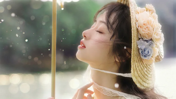 꽃 장식이 달린 밀짚모자를 쓴 성인 여성이 부드러운 보케 빛 속에서 눈을 감고 있습니다.
