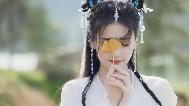흰색 전통 의상을 입은 여성이 야외에서 눈을 감고 금빛 잎을 들고 있는 부드러운 배경의 인물 사진이다.