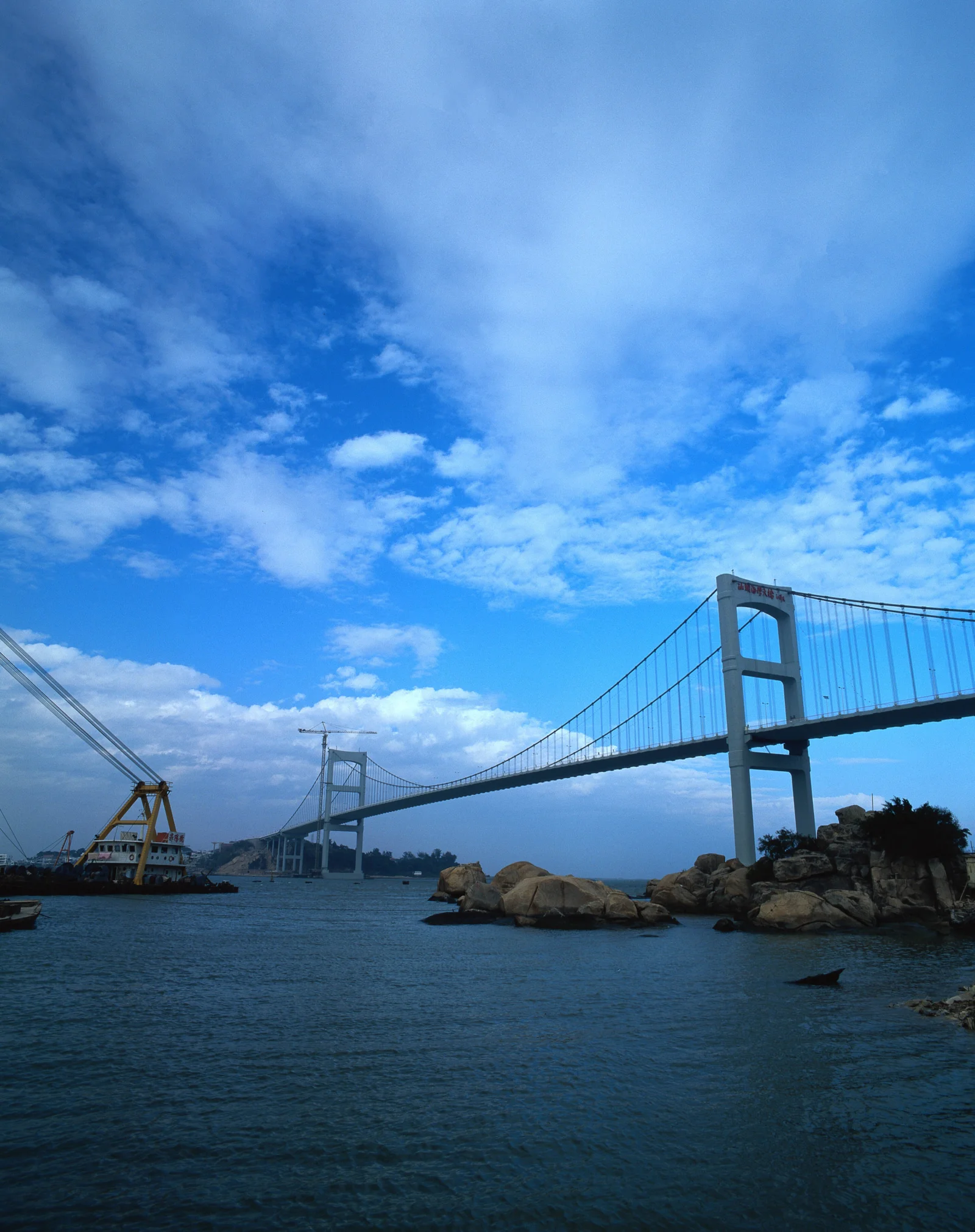 一座悬索桥横跨海面，蓝天、云层和岸边岩石构成画面主体。