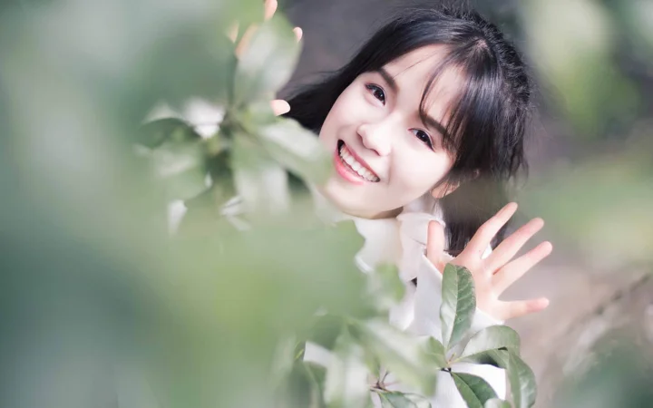 一名微笑的女性站在绿叶间，抬手向镜头挥手。
