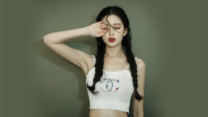 一名留着双辫的女性在灰绿色背景前闭眼摆出肖像姿势。