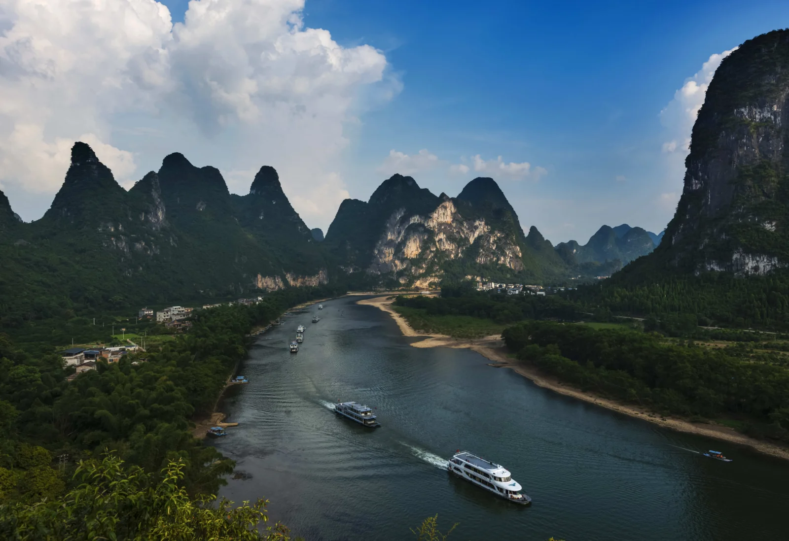 漓江穿过群山与绿树，两艘游船行驶在河面上，天空飘着大片白云。