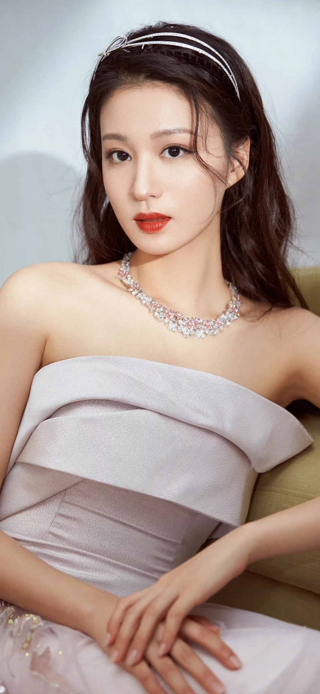 一位长发女性身穿浅色礼服，佩戴项链并坐在室内沙发上。