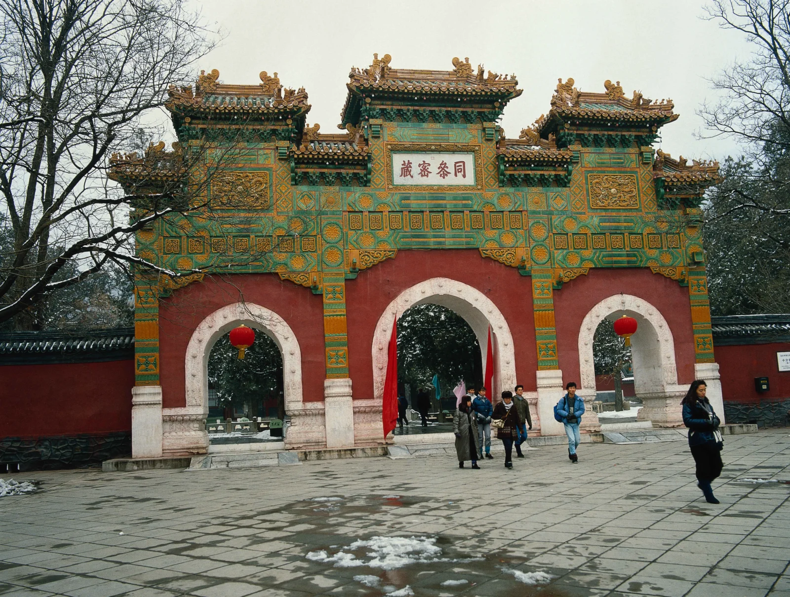 一座色彩鲜艳的中式古建筑牌楼矗立在铺石庭院中，周围可见树木和少量积雪。