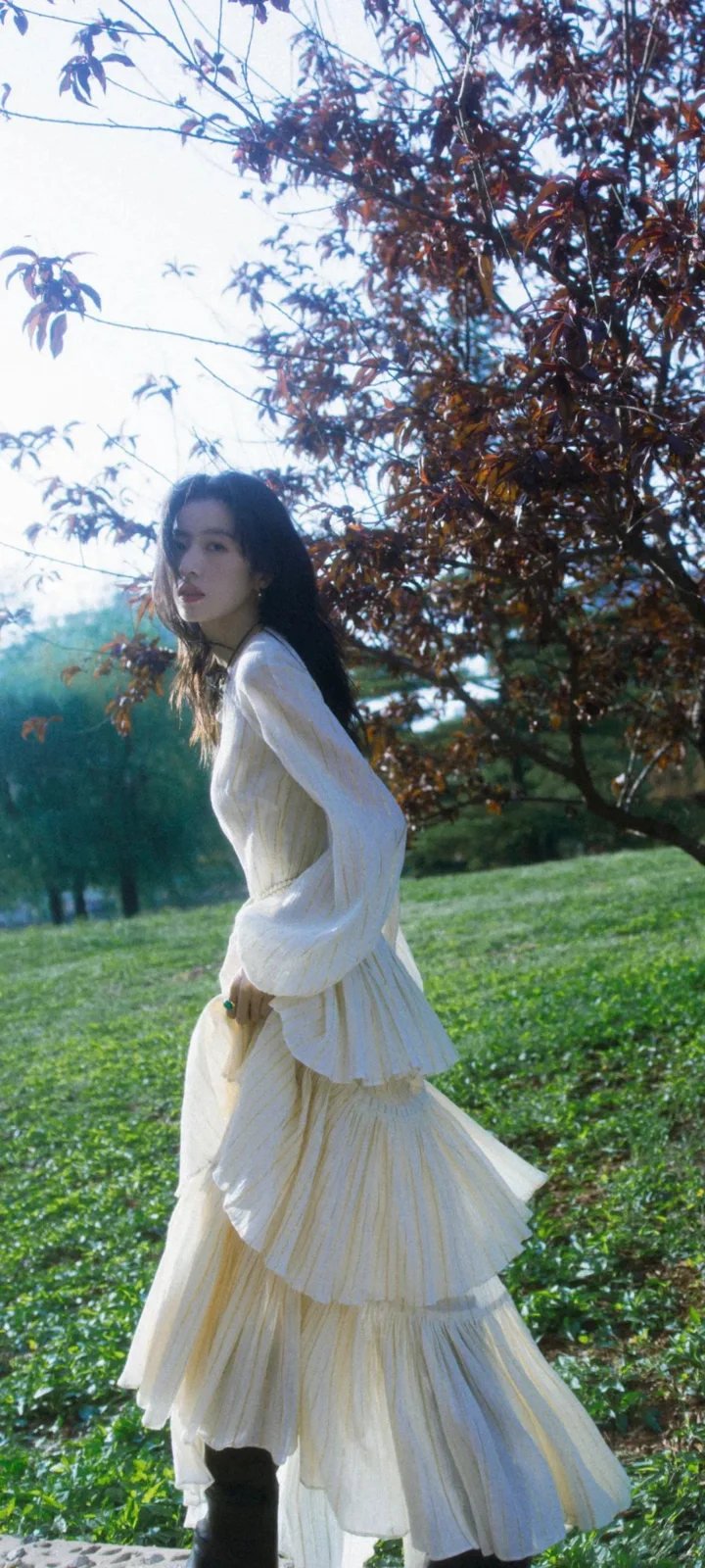 一名身穿白色长裙的女子站在树木与草地之间的户外花园中。
