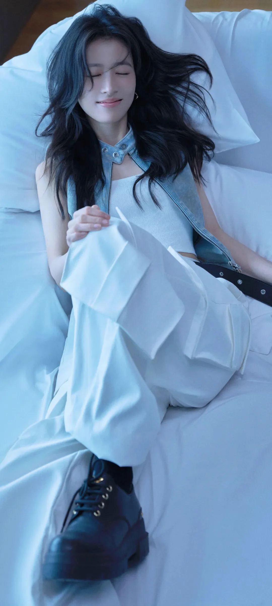 긴 머리의 여성이 흰색 드레스를 입고 흰 침대에 누워 검은 부츠를 신고 있다.