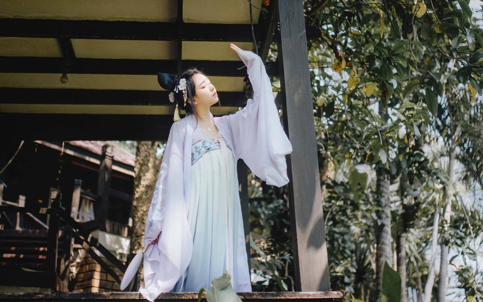 밝은 색상의 전통 의상을 입은 여성이 나무로 된 구조물 옆에 나무들로 둘러싸여 서 있습니다.