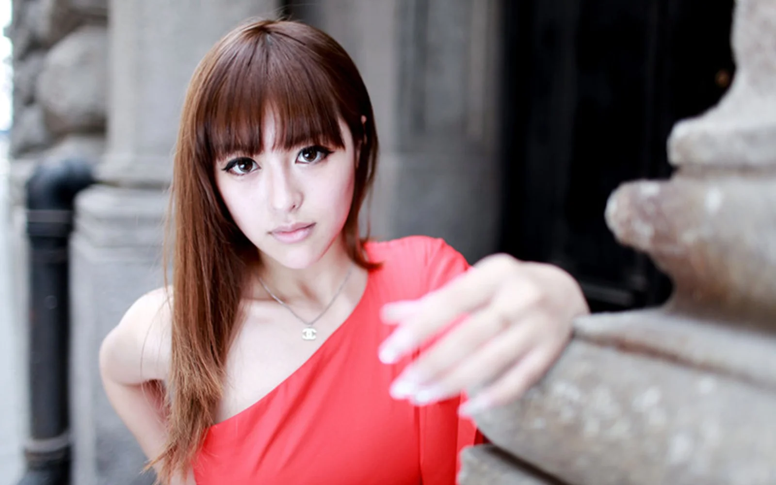 一名棕色长发女子穿着红色上衣，在石质建筑前看向镜头。