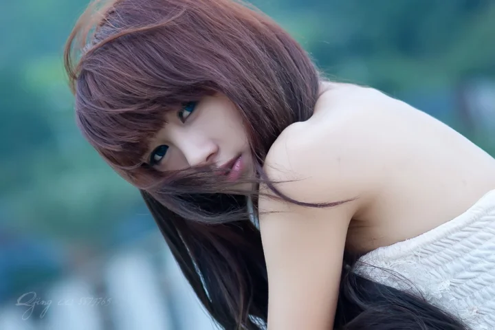 一名长发女子穿着白色纹理上衣，俯身凝视镜头，背景呈现蓝绿色的虚化景象。