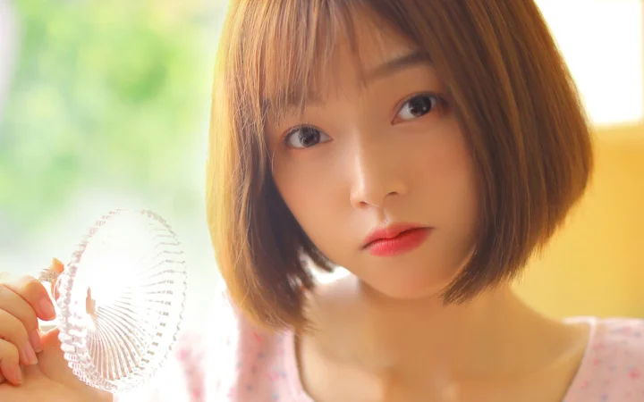 一名短发女性手持白色扇子面对镜头，背景呈现柔和的绿色与暖黄色光影。