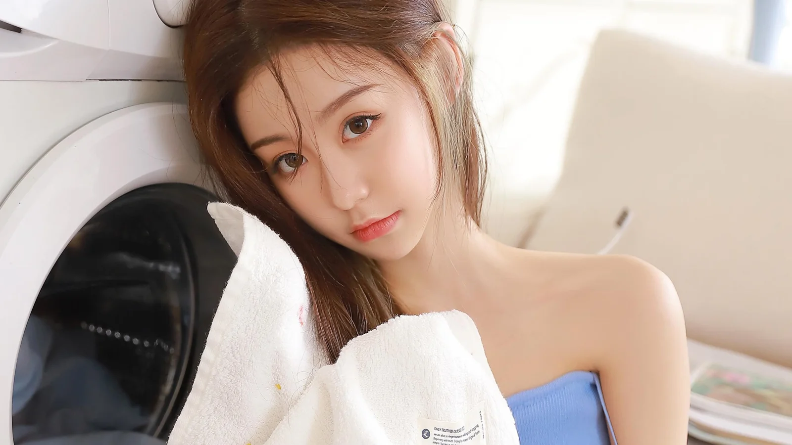 갈색 긴 머리의 여성이 파란색 상의를 입고 세탁기 옆에서 흰 수건을 들고 있습니다.