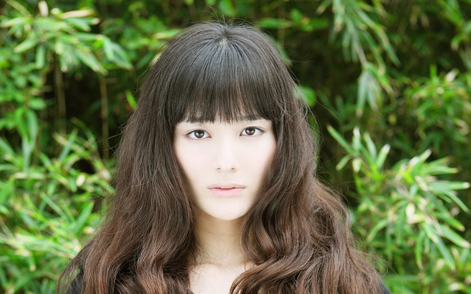 一名长发女子站在竹林和绿色植物前，面向镜头。