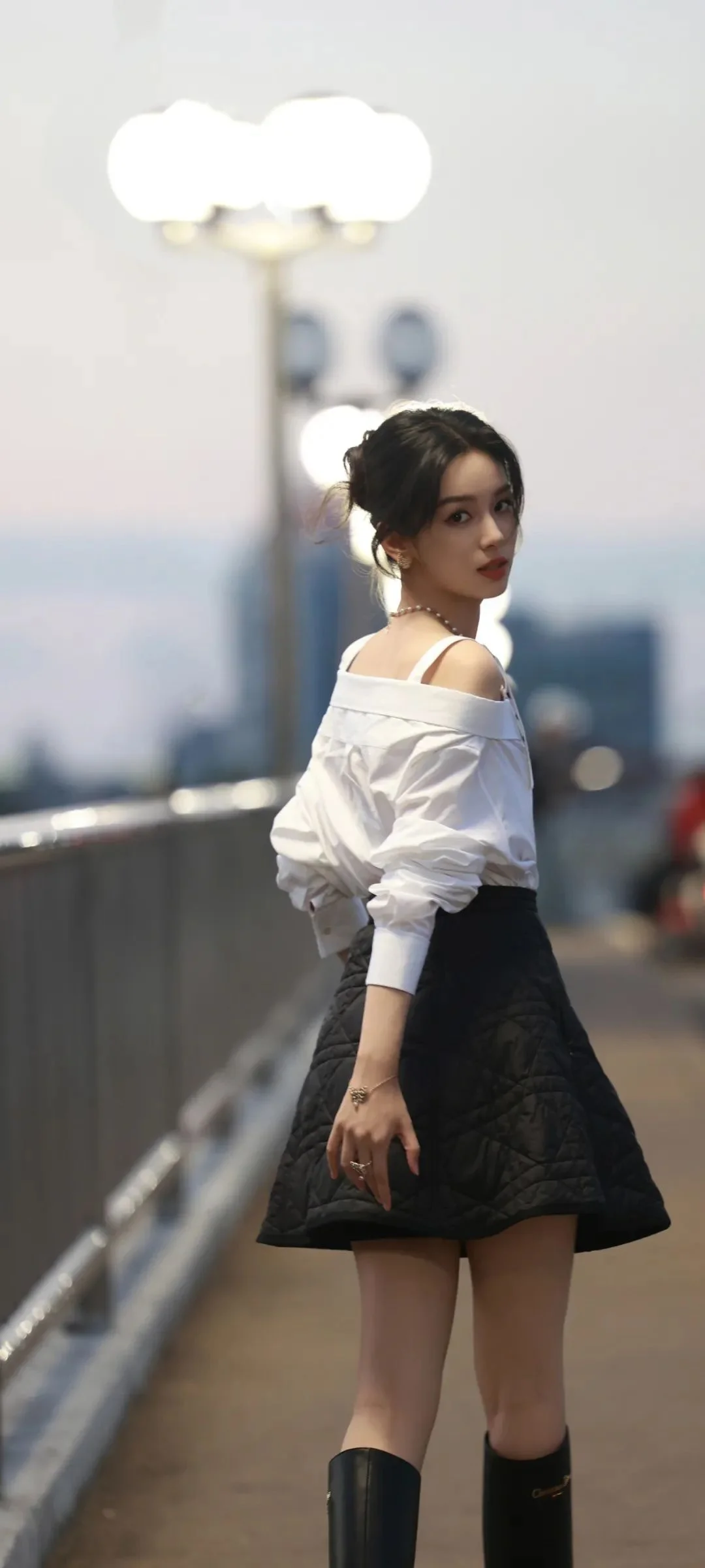 一名女子穿着白色露肩衬衫、黑色短裙和长靴，站在傍晚的城市人行桥上回眸。