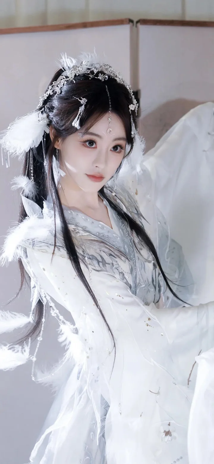 白い伝統衣装を着て羽飾りを身につけた女性が室内に立っています。