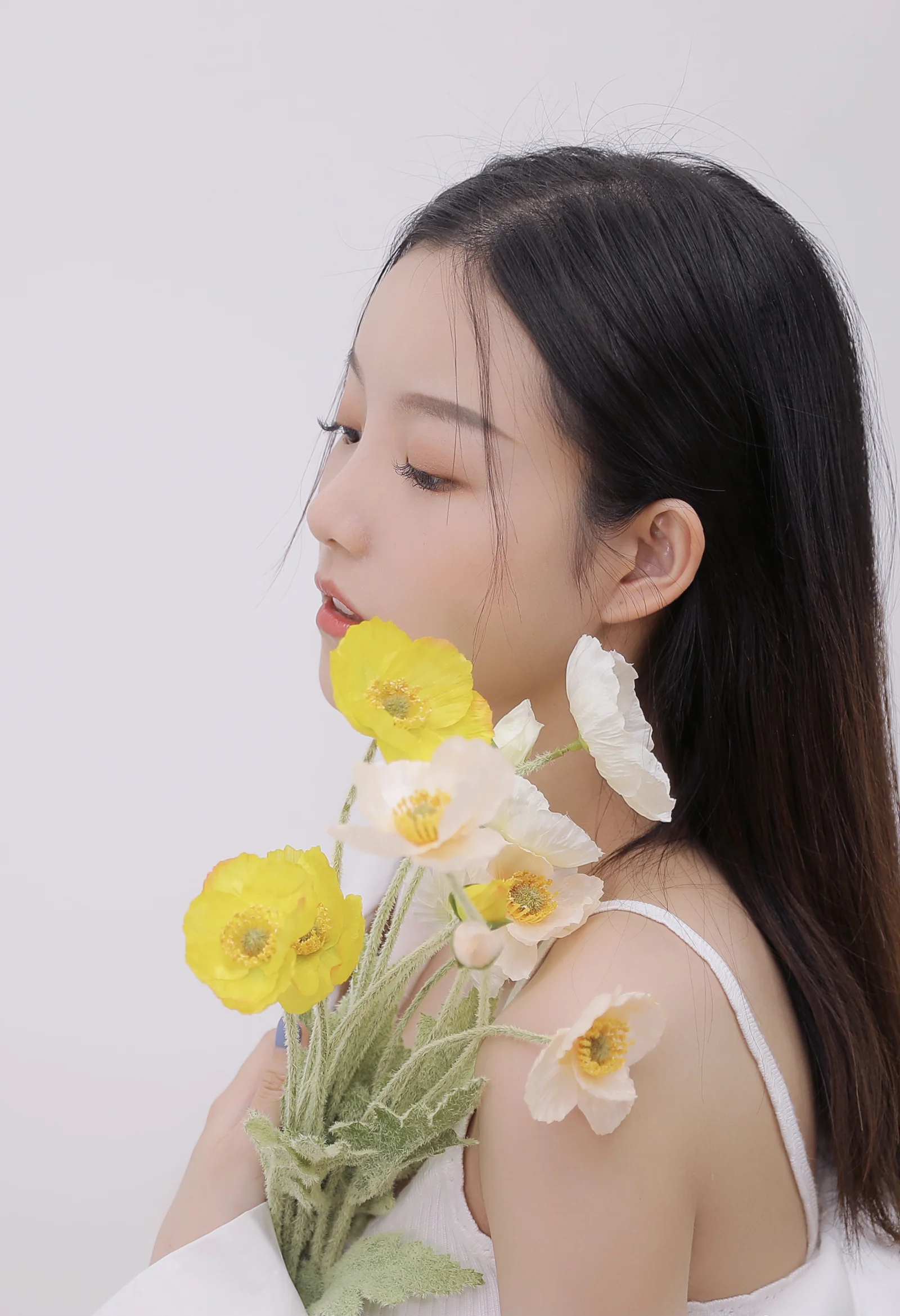 一名长发女子侧身手持黄色和白色鲜花，背景为浅灰色。