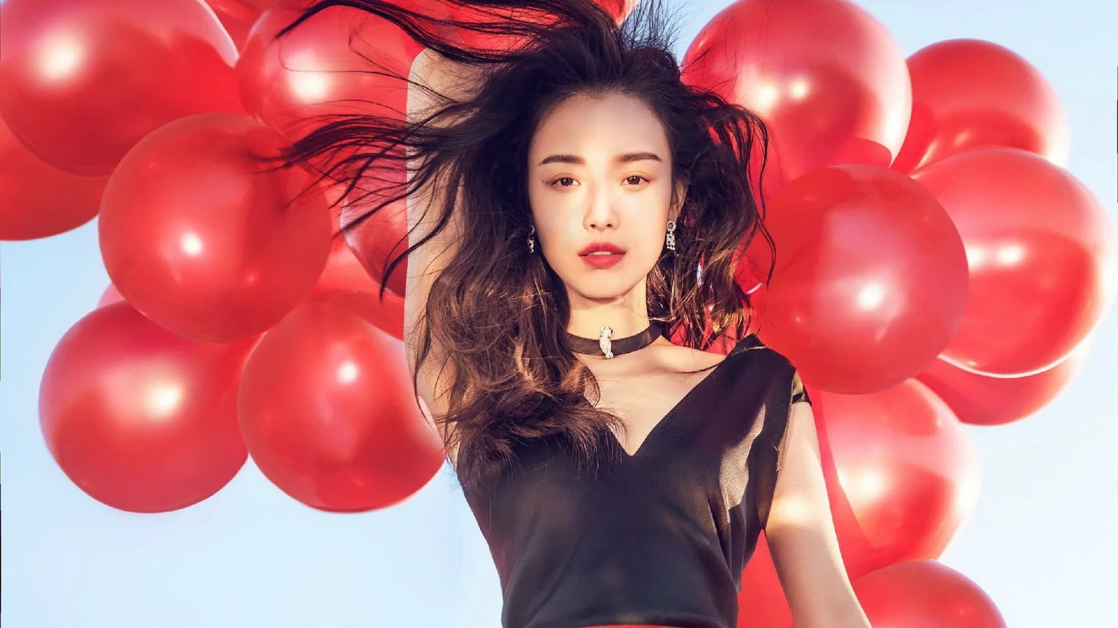 一名身穿黑色服装的女子站在一群红色气球前，头发随风飘动。
