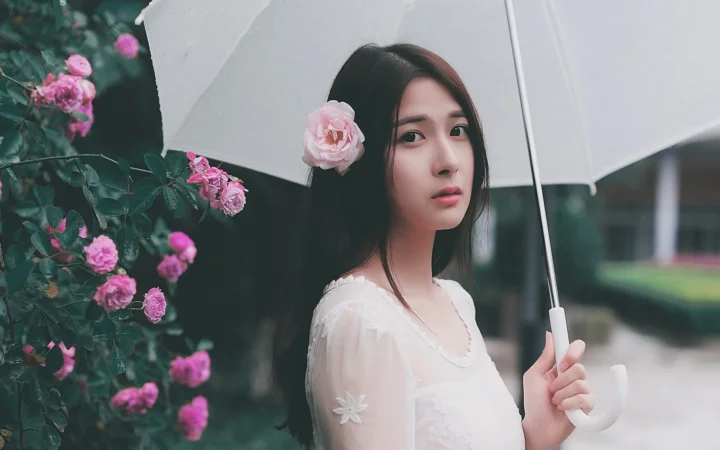 一名年轻女子手持白色雨伞站在盛开的粉色玫瑰旁。