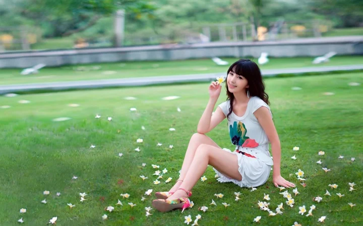 一名女子坐在铺满白色小花的绿色草地上，手持一朵黄色小花。