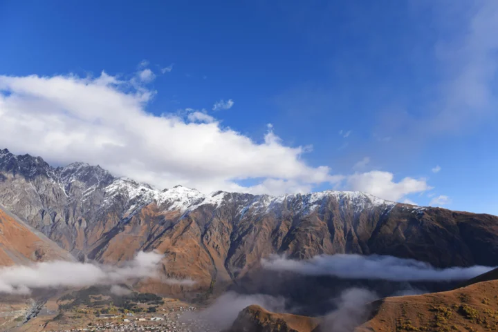 巍峨的雪山横跨画面，云雾缭绕在山腰，远处山脚可见一片村落。