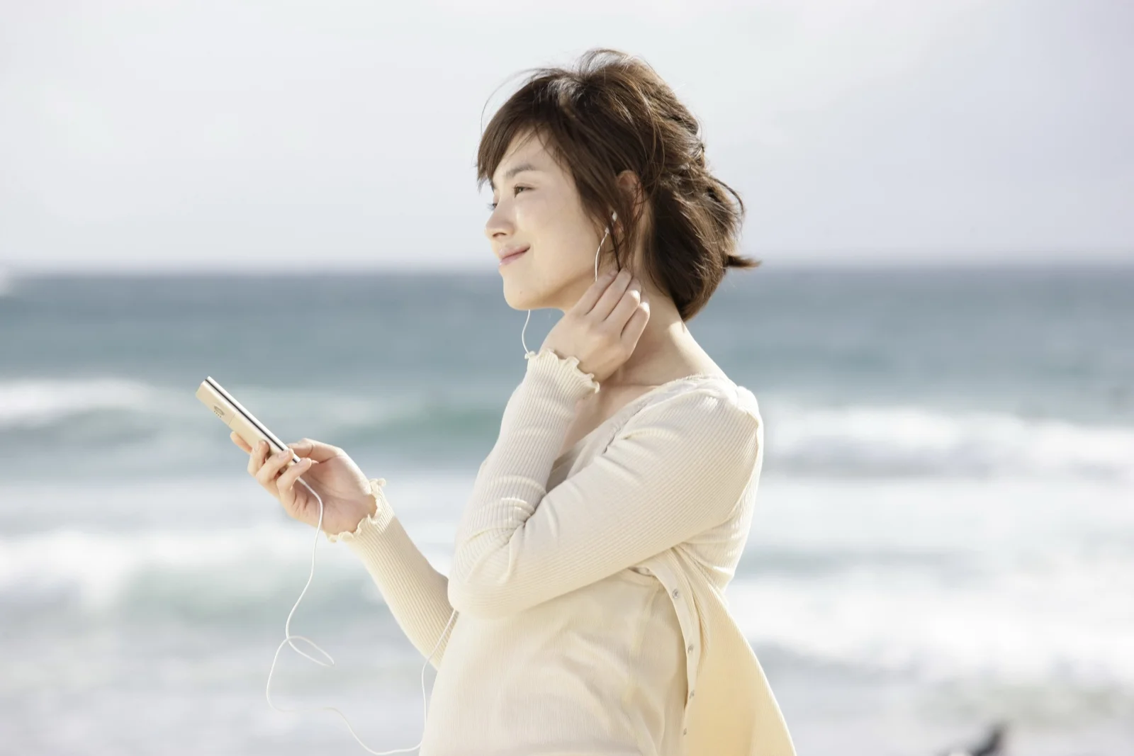 한 여성이 바닷가에서 스마트폰을 들고 이어폰으로 음악을 들으며 바다를 바라보고 있다.