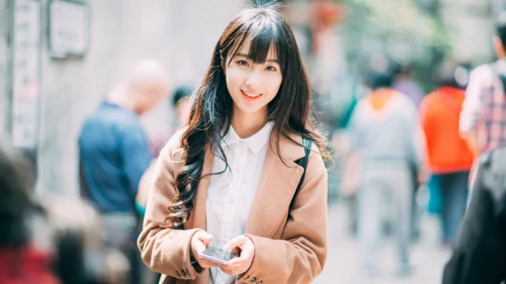 長い髪の女性が茶色のコートと白いシャツを着て、スマートフォンを手に街角に立っており、背景の人々はぼやけている。