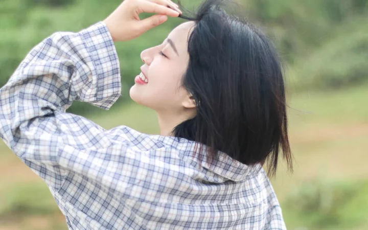 一名穿格纹衬衫的成年女子在户外微笑并抬手整理头发。