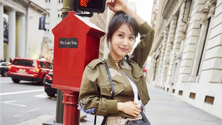 성인 여성이 건물과 차량, 보도가 보이는 도시 거리에서 빨간 우체통 옆에 서 있습니다.