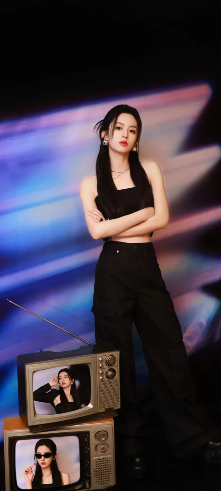 검은색 옷을 입은 성인 여성이 화려한 빛의 배경 앞에 서 있고 화면 아래에는 복고풍 텔레비전 두 대가 놓여 있다.