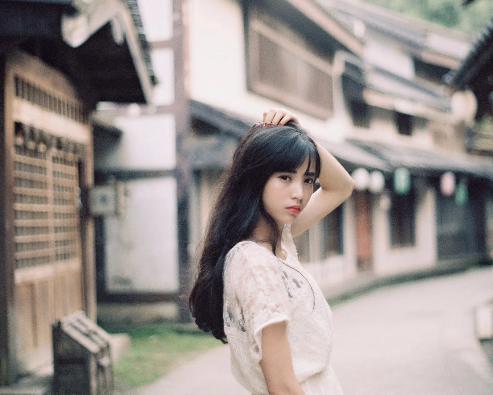 一名长发女子站在老街巷中，抬手触碰头发，背景是虚化的建筑与街道。