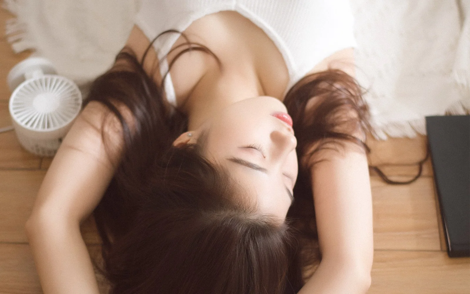 一名長髮女子躺在木地板上，頭部側向一旁，周圍可見白色織物和小型風扇。