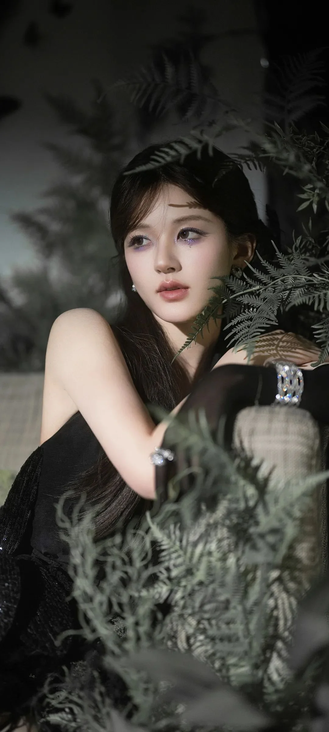 一位女性身穿黑色服装，置身蕨叶环绕的暗色背景中。