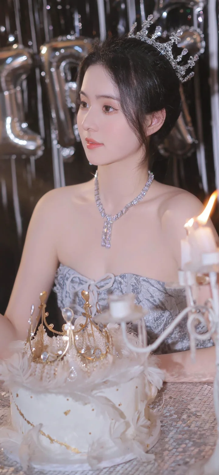 一位戴着水晶皇冠和项链的女子身穿礼服坐在烛光前。