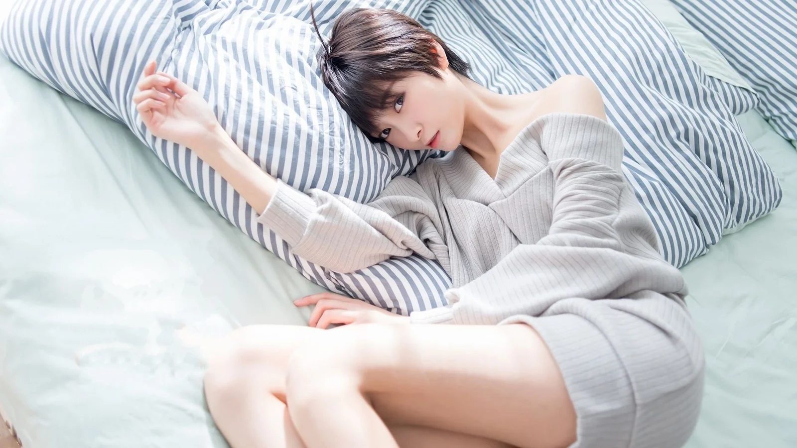 淡い色のニットを着た成人女性が、ストライプ柄の枕に頭を預けてベッドに横たわっています。