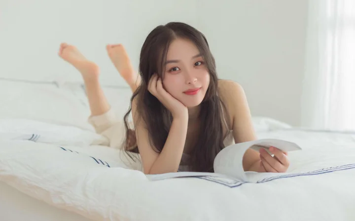 一名成年女性趴在床上阅读书籍，画面明亮柔和。
