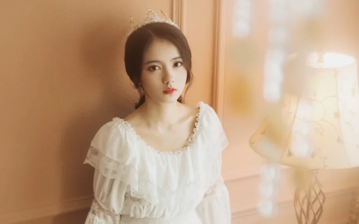 작은 티아라를 쓰고 흰 드레스를 입은 여성이 따뜻한 조명 아래 벽과 테이블 램프 근처에 앉아 있습니다.