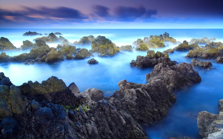 长曝光效果呈现出蓝色海水环绕嶙峋岩石的海岸景观，远处天空被云层覆盖。