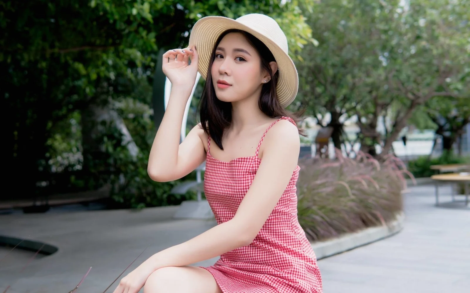 밀짚모자와 분홍색 깅엄 체크 원피스를 입은 성인 여성이 녹음이 우거진 야외 보도 옆에 앉아 있습니다.