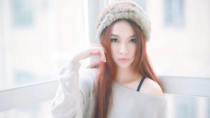 ニット帽をかぶった赤毛の女性が、明るい窓辺で淡い色のゆったりしたトップスを着て座っています。