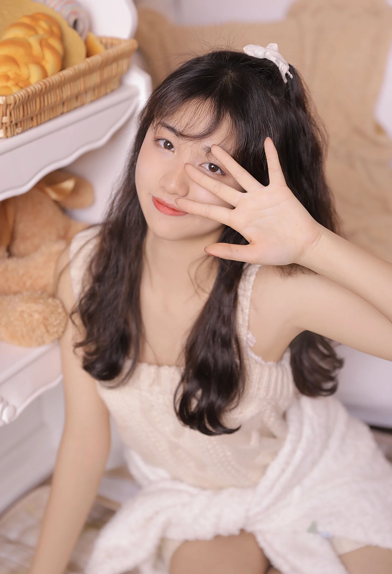 一名长发女性身穿白色连衣裙，在室内抬手遮住部分面部面对镜头。