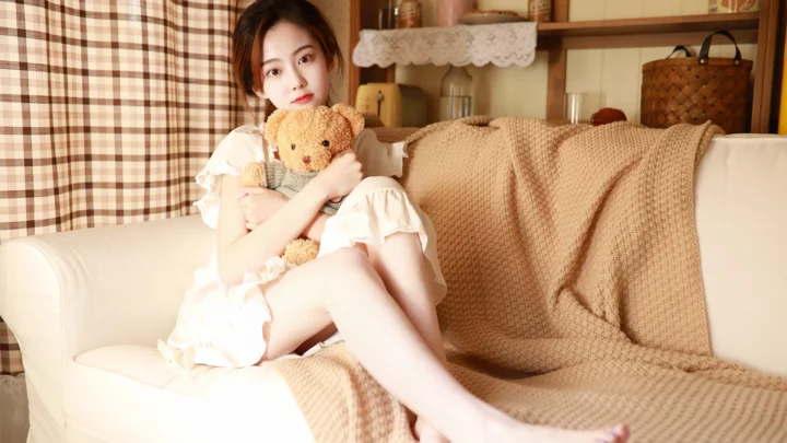 一名女性穿着白色连衣裙坐在沙发上抱着泰迪熊，身后是格纹窗帘和室内置物架。
