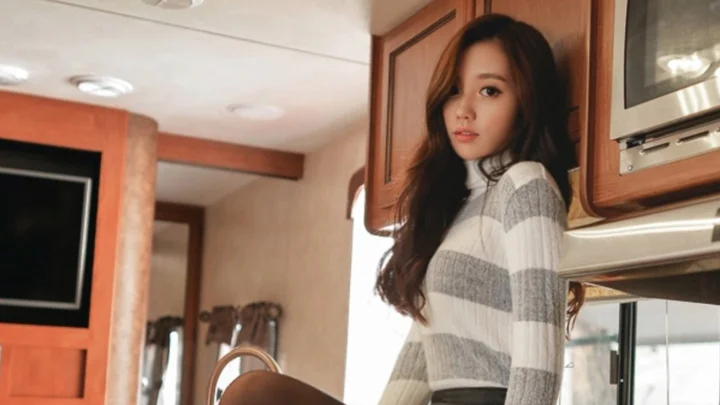 긴 머리의 여성이 나무 수납장과 주방 설비 옆 실내에 서 있습니다.