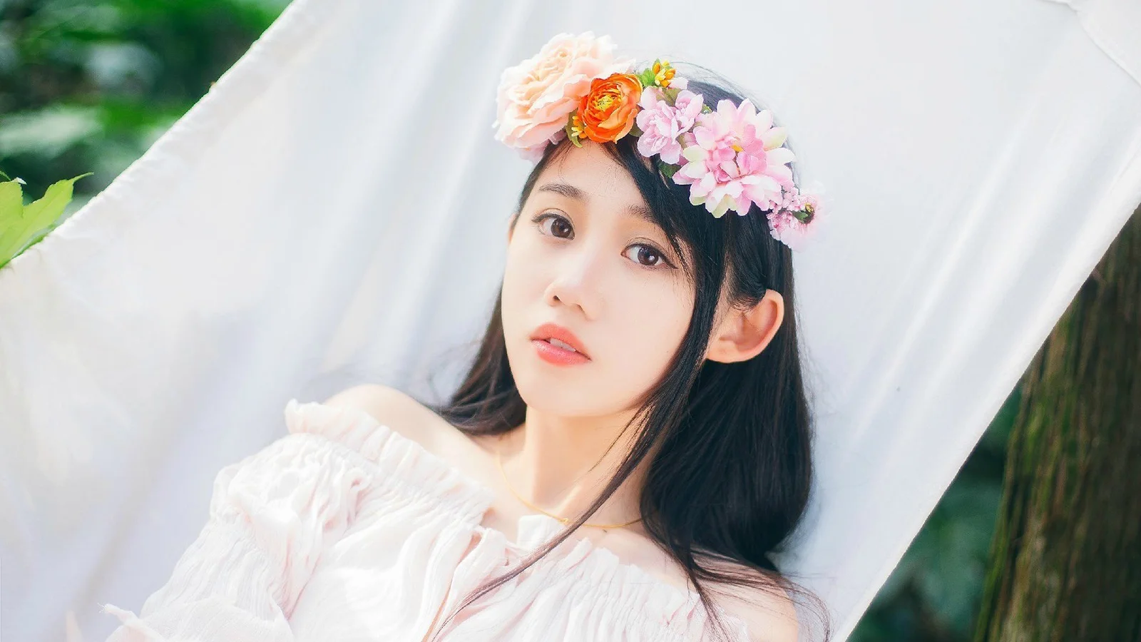 花冠を身につけた女性が白い布のそばに寄りかかり、背景には緑の植物が見えます。