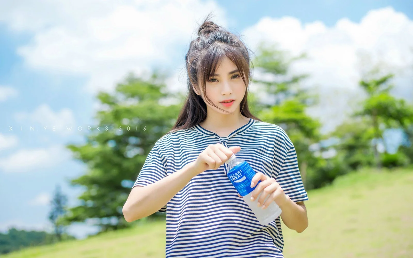 一名长发女性在蓝天绿地间手持透明水瓶。