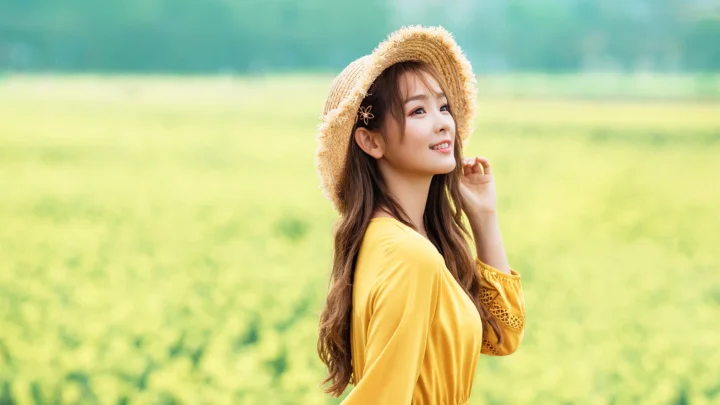 一名戴草帽、穿黄色连衣裙的女子站在明亮的绿色田野中。