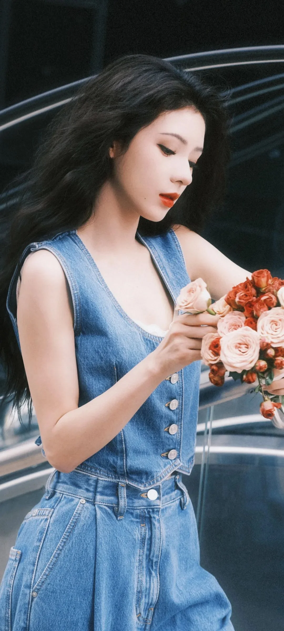 파란 데님 옷을 입은 여성이 어두운 배경 앞에서 분홍색과 빨간색 꽃다발을 들고 있습니다.