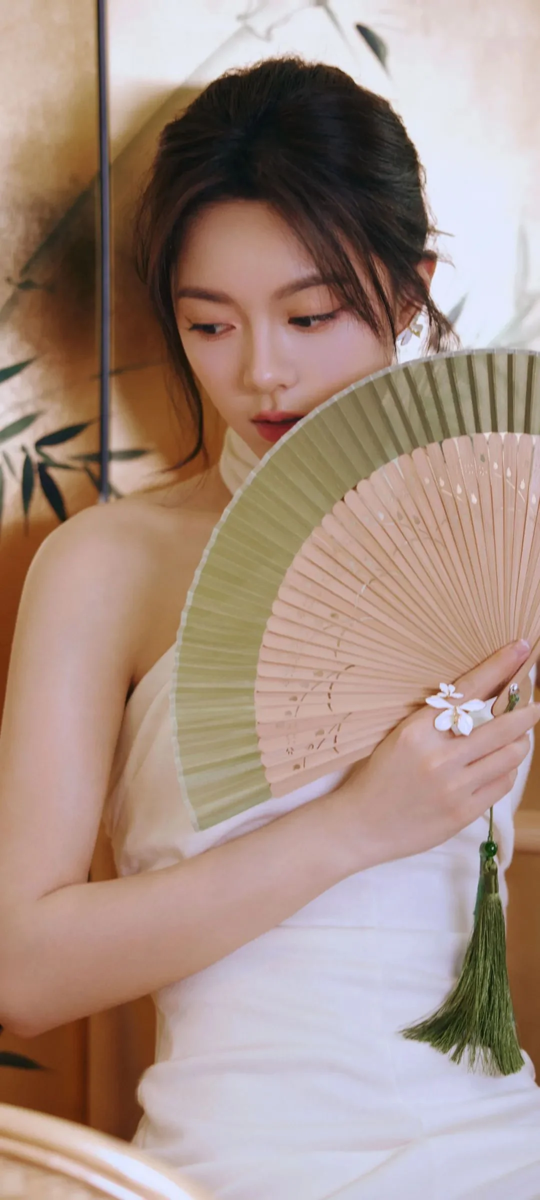 一名身穿白色服装的女性手持折扇，室内暖色背景下侧目低头。