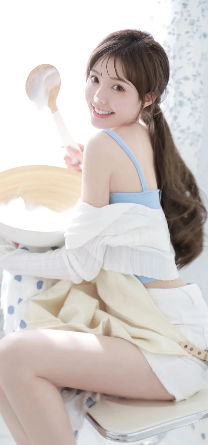 一名长发女性回头微笑，手持勺子坐在室内浅色背景前。